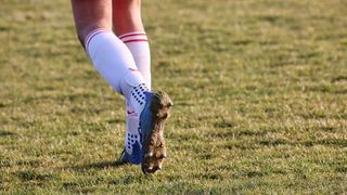 A soccer player is standing on a field, their foot wearing a blue and white soccer cleat with mud. The player has red stripes on their white socks.
