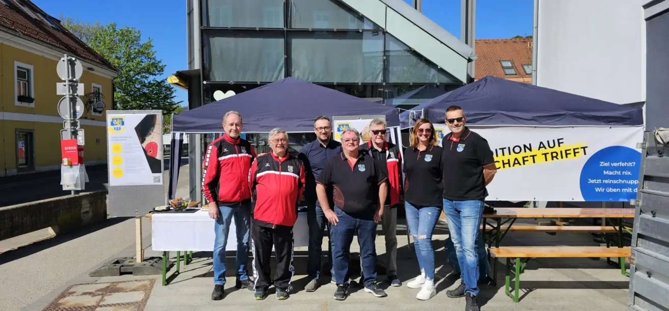 A group of people wearing black and red jackets stand in front of a tent and a building. A table with food is behind them.