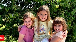 Three young girls smile at the camera, with one wearing a pink shirt, one in a yellow shirt with a deer design, and the third in a white shirt. They are standing close together in front of green foliage.