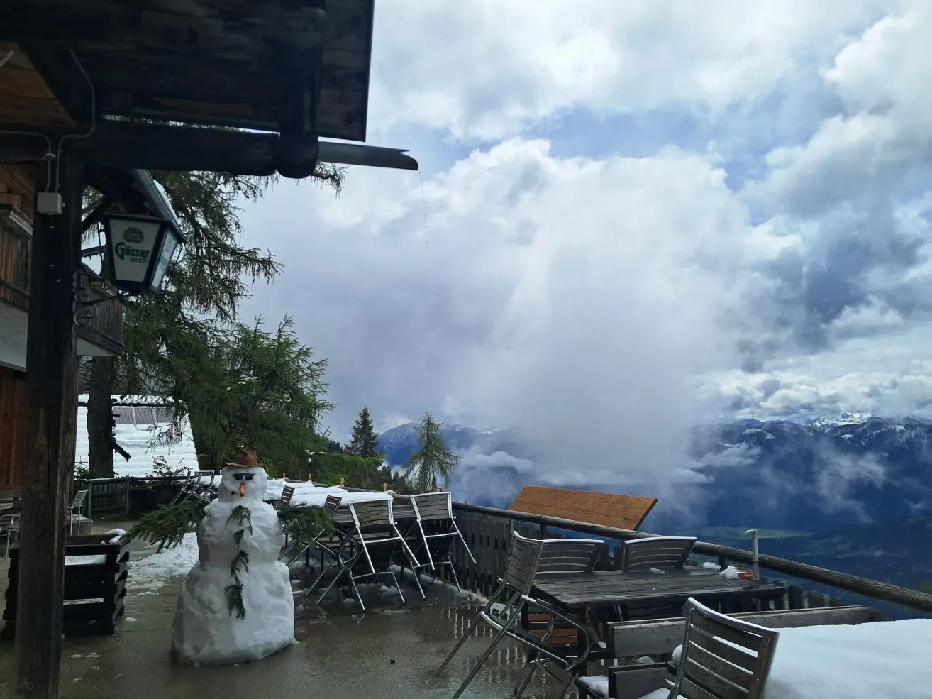 Eine verschneite Landschaft zeigt einen Schneemann auf der Terrasse, umgeben von Tischen, Stühlen und einem Baum. Dahinter sind Berge mit Wolken bedeckt.