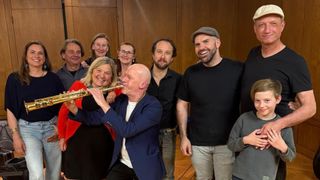 A group of musicians, including a man playing a flute, are smiling and posing for a photo in a room.