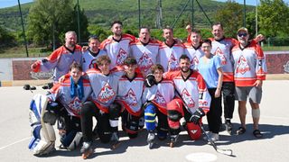Eine Hockeymannschaft posiert für ein Foto in ihren Teamtrikots. Sie stehen und knien auf einem Betonuntergrund mit einem Zaun im Hintergrund.