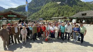 A group of people is posing for a photo on a brick pathway in front of a mountain. There are several people wearing hats and glasses. There are two streetlights on the right.