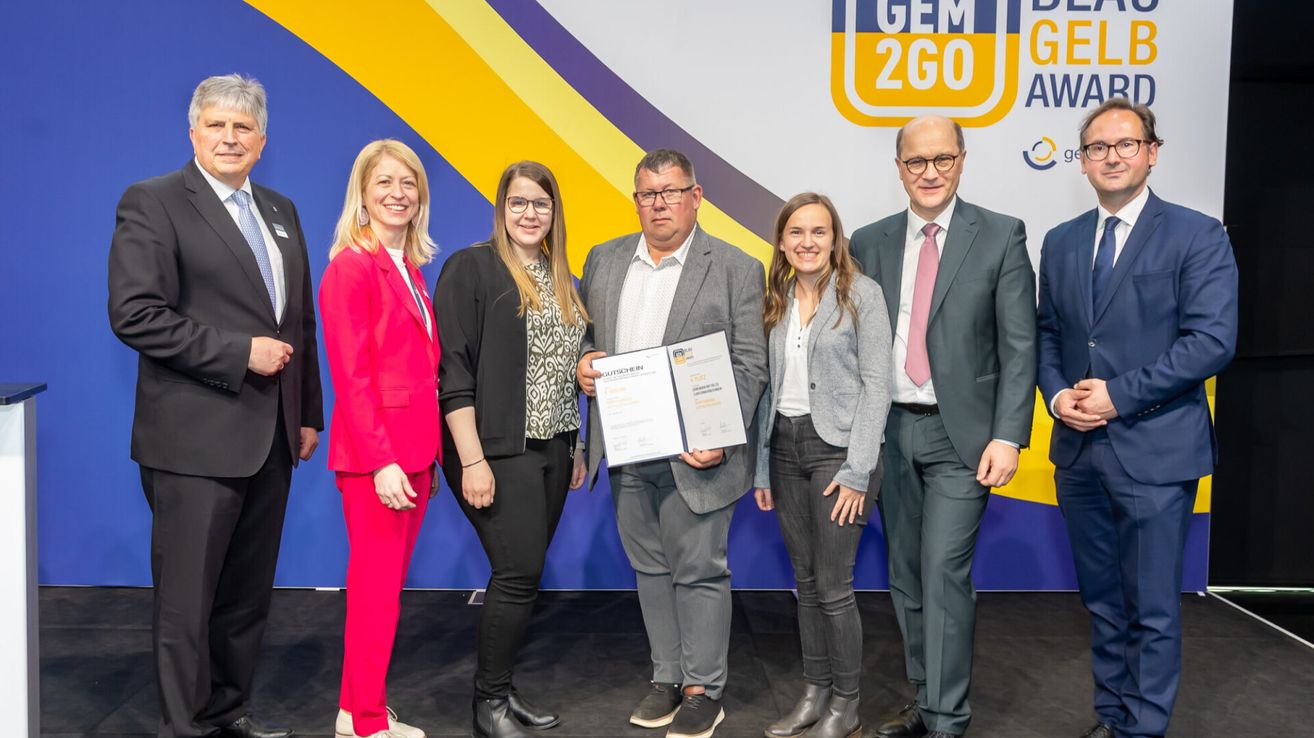A group of people stands on a stage holding a certificate. Four of them are smiling, and one man looks serious. Behind them, a banner reads GEM 2GO.