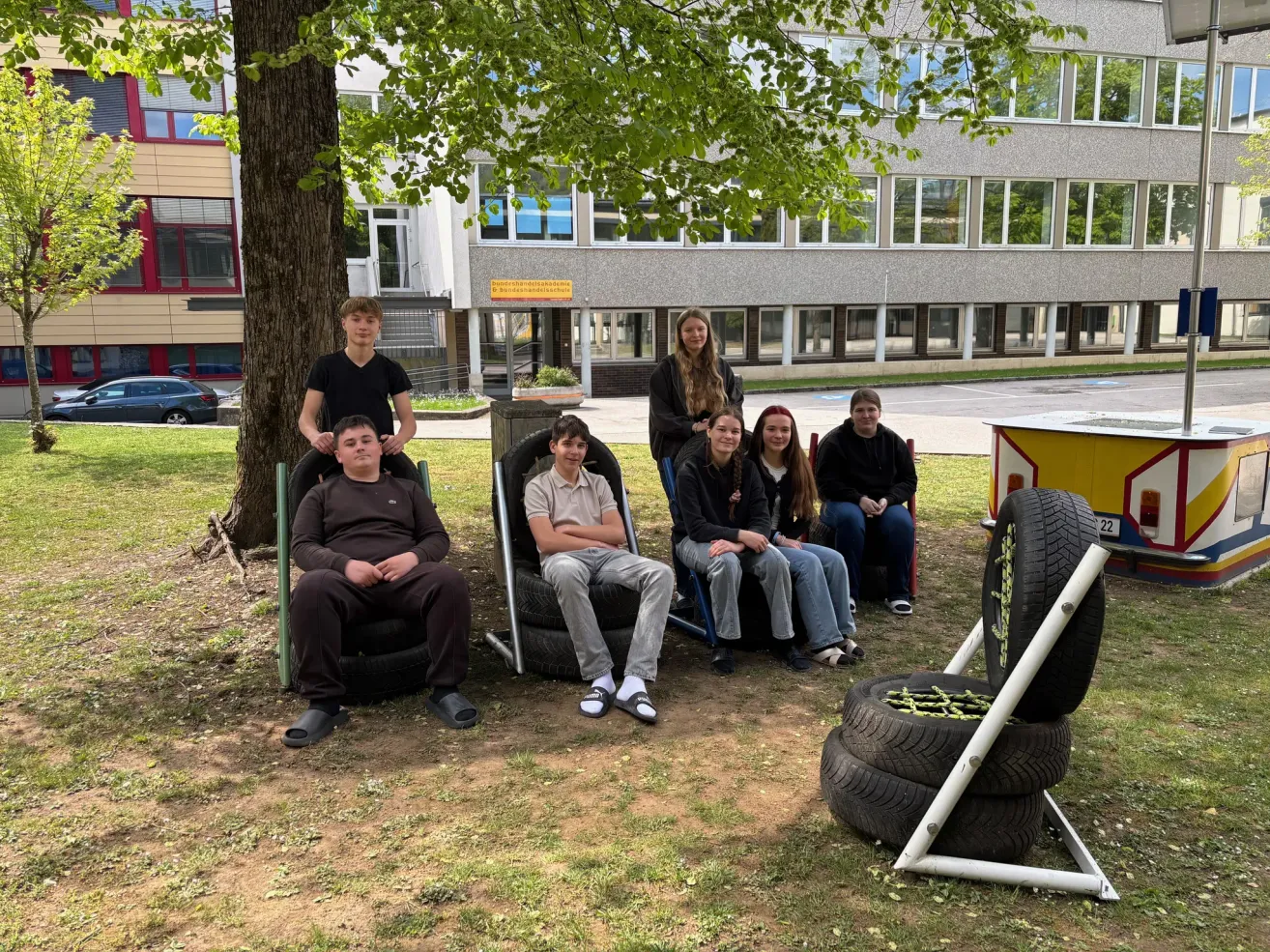 Seven young people sit on tires in a park with a building in the background. Three sit in chairs, and two are leaning against a tree. A yellow stand with tires is in the foreground.