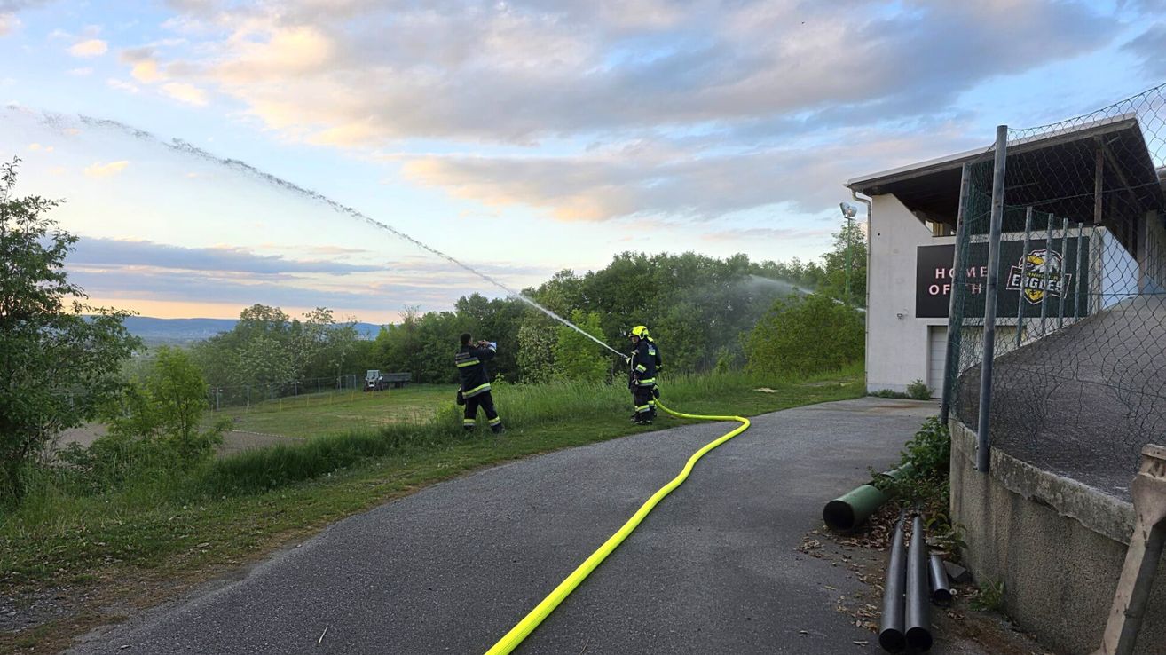 Two firefighters are using a water hose to spray water outside. They are on a paved path near a building.