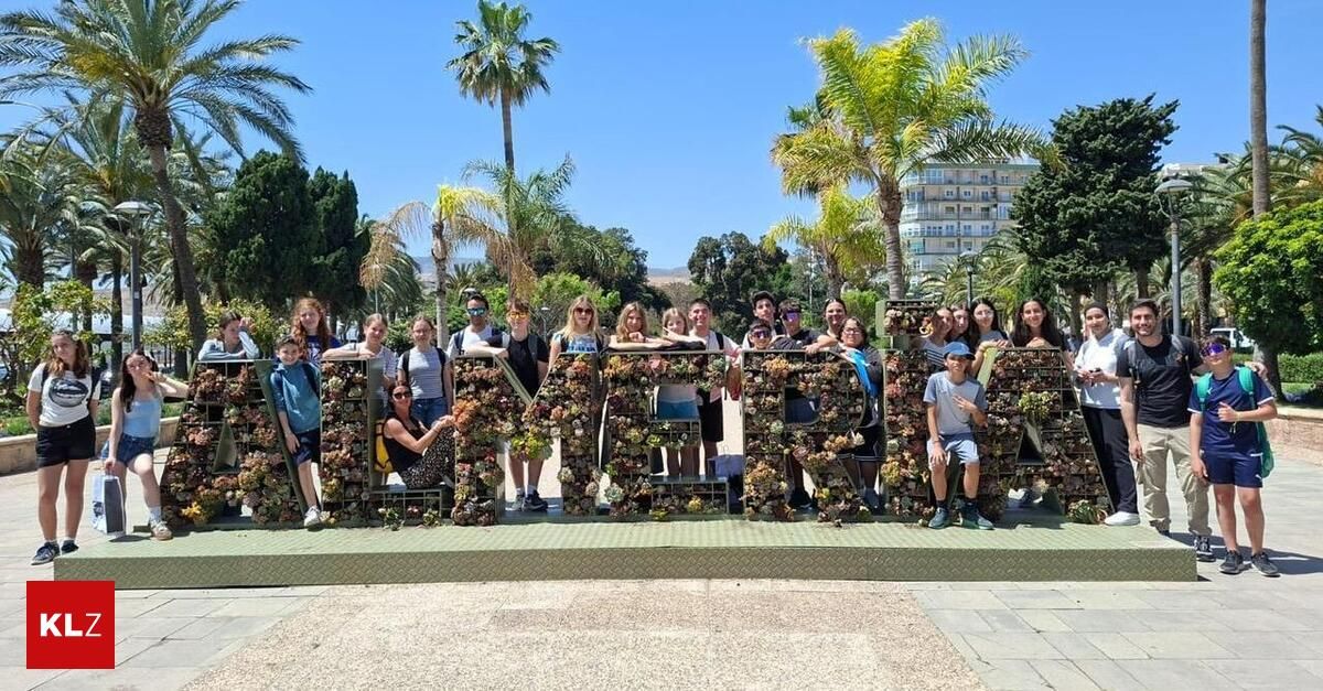A group of people pose for a photo in front of a large letter display in a park. They are wearing casual clothing and some are wearing sunglasses. The backdrop features palm trees and a building.