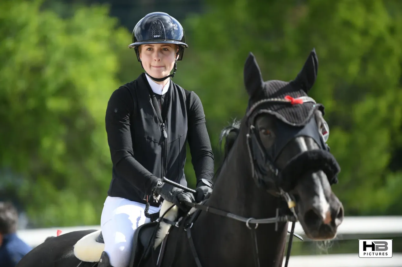Eine Frau in Reitausrüstung reitet ein schwarzes Pferd auf einem grünen Feld. Sie trägt einen Helm, Handschuhe und hält die Zügel mit selbstsicherer Haltung.