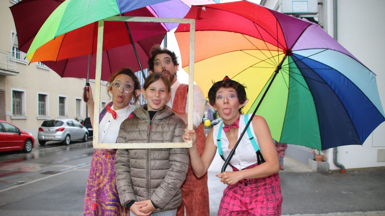 Four individuals dressed in costumes pose with umbrellas. Two of them have their tongues out, while one holds a picture frame. They appear to be having fun.