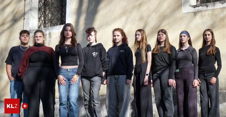 Seven young women and one man in hoodies stand in a row outside a beige wall, likely posing for a photograph.