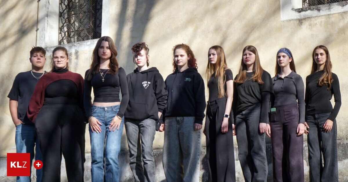 Seven young women and one man in hoodies stand in a row outside a beige wall, likely posing for a photograph.