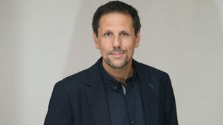 Ein Mann mit Bart und Oberlippenbart steht lächelnd für ein Foto. Er trägt ein dunkelblaues Hemd und einen Blazer. Er befindet sich wahrscheinlich in einem Büro.