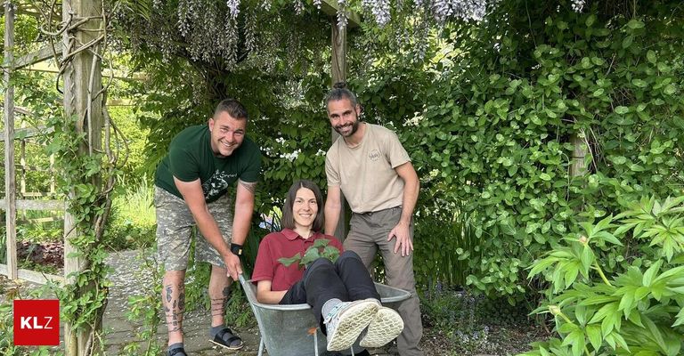 Drei Menschen in einem Garten. Eine Frau sitzt in einer Schubkarre und lächelt. Zwei Männer stehen hinter ihr, lächeln und halten an der Schubkarre.