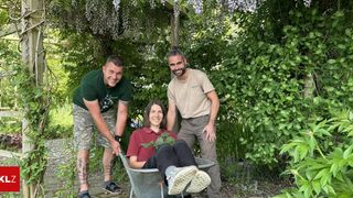 Three people in a garden. A woman sits in a wheelbarrow, smiling. Two men stand behind her, smiling and holding onto the wheelbarrow.