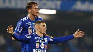 Two soccer players in blue uniforms celebrate. One has a KARO shirt, and the other has multiple sponsor logos on his jersey.