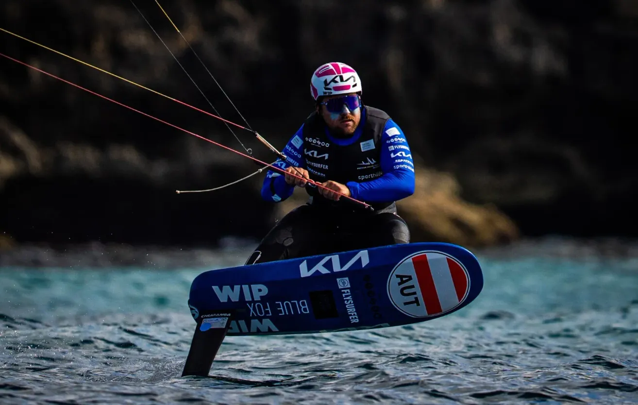 A windsurfer glides on the water, holding onto the ropes, with a blue board and a blue outfit. The board has a white logo with red letters and a round emblem.