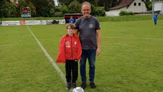 A man and a young boy are standing on a soccer field with a soccer ball. Trees and a house are in the background.
