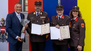 Three men in military uniforms hold certificates in front of a flag. One holds a bouquet.