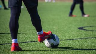 A soccer player with red shoes is kicking a ball on a field. Another player is blurry in the background.