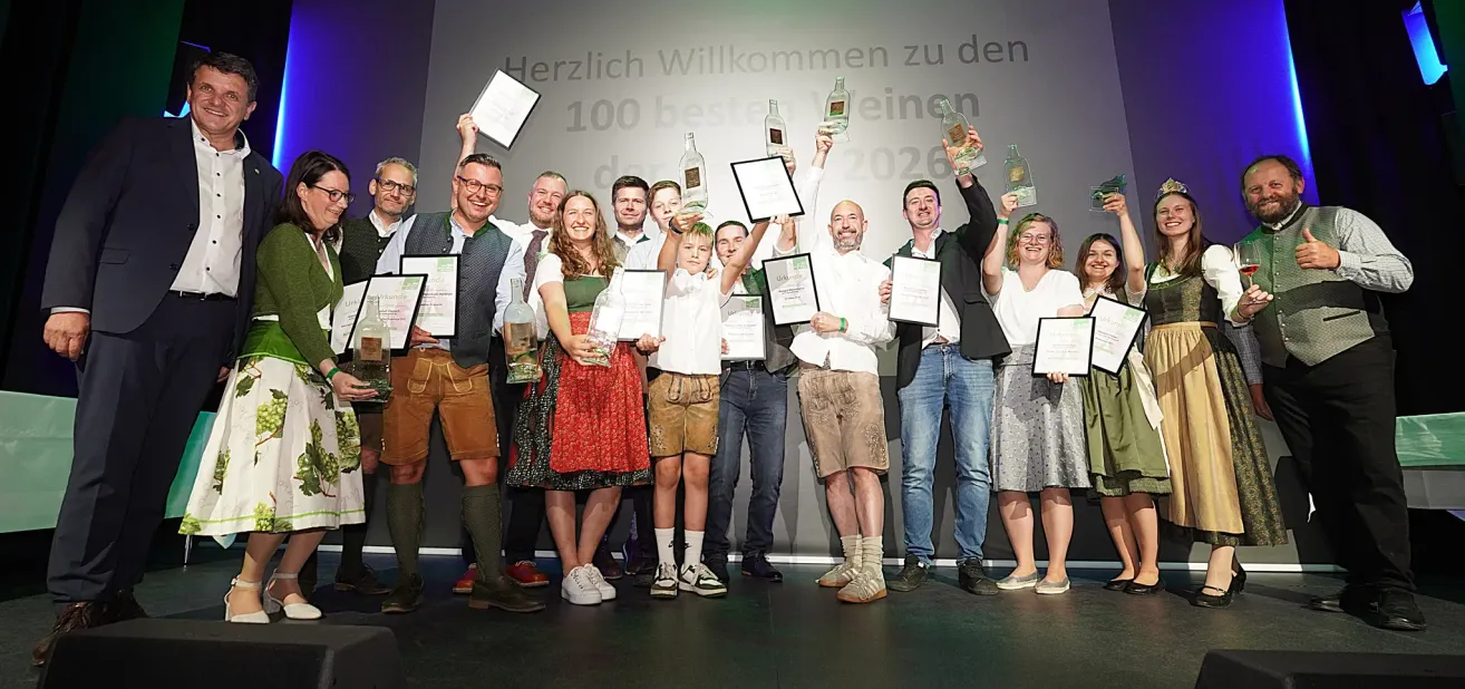 A group of adults and children are posing for a photo on a stage. They hold bottles and certificates. The wall behind them has a white backdrop with text.