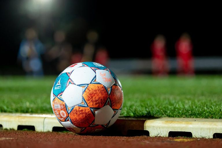 A soccer ball with a blue, white, and orange design sits on a grassy field. Behind it, people in red jerseys stand.