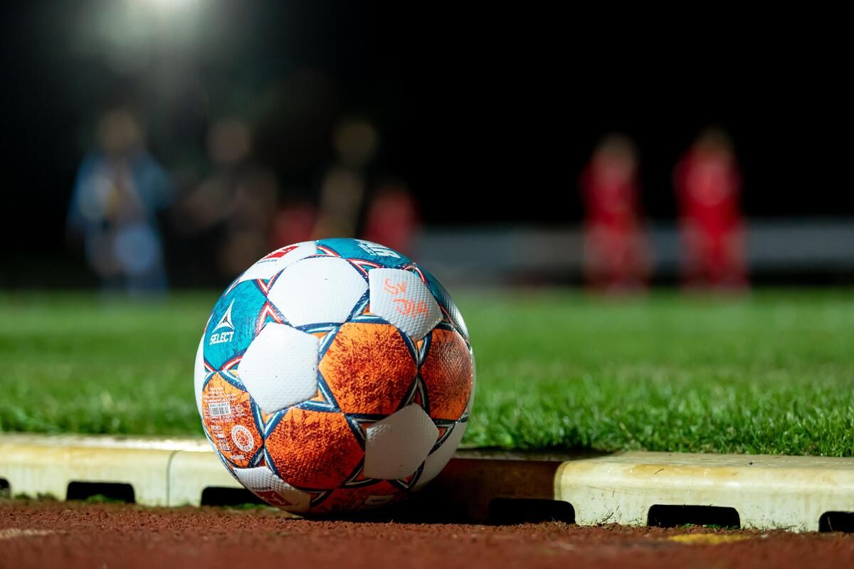 A soccer ball with a blue, white, and orange design sits on a grassy field. Behind it, people in red jerseys stand.