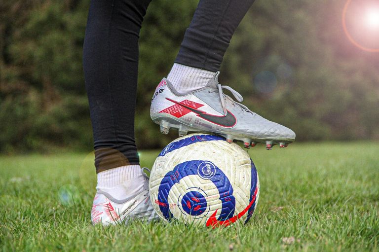 A soccer player stands on a soccer ball, wearing Nike cleats. The ball has a blue and red design with a logo. The background is a blurry green landscape.
