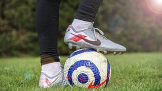 A soccer player stands on a soccer ball, wearing Nike cleats. The ball has a blue and red design with a logo. The background is a blurry green landscape.