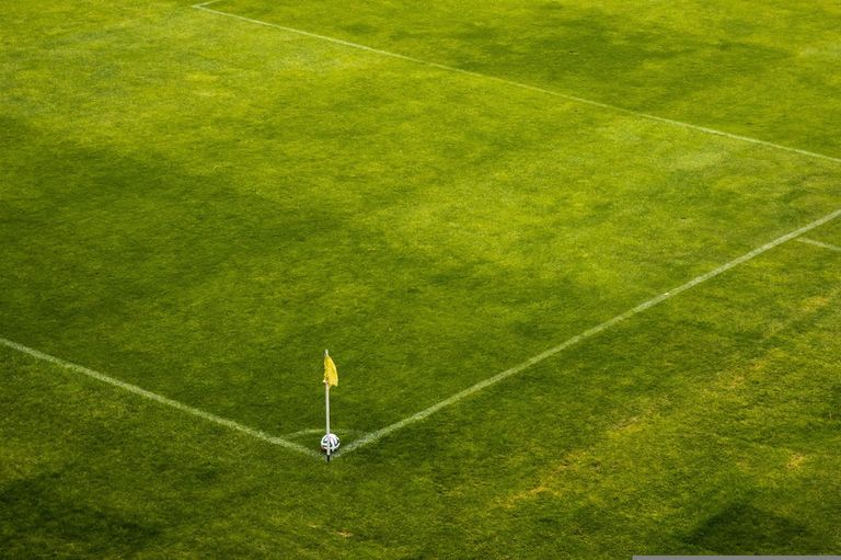 An empty soccer field with green grass and white lines. A yellow flag and a soccer ball are near the corner.