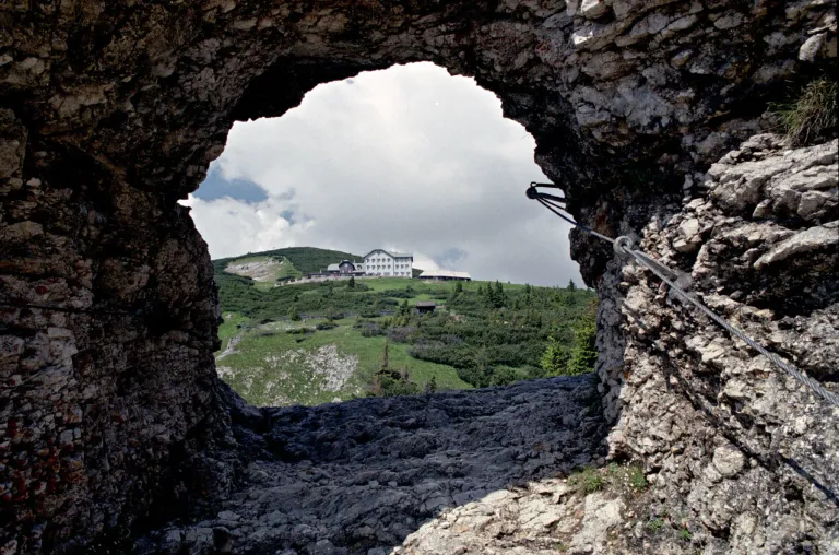 Ein Blick durch ein Loch in einem Felsen zeigt ein großes weißes Gebäude auf einem grünen Hügel mit einem bewölkten Himmel im Hintergrund.