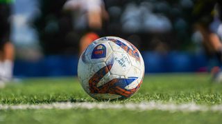 A soccer ball is on the field. It has blue and orange colors with white writing. There is a blurred background.