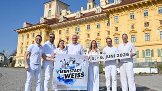 Eine Gruppe Erwachsener, die in Weiß gekleidet sind, posieren mit Schildern vor einem gelben Gebäude am 5. und 6. Juni 2026.
