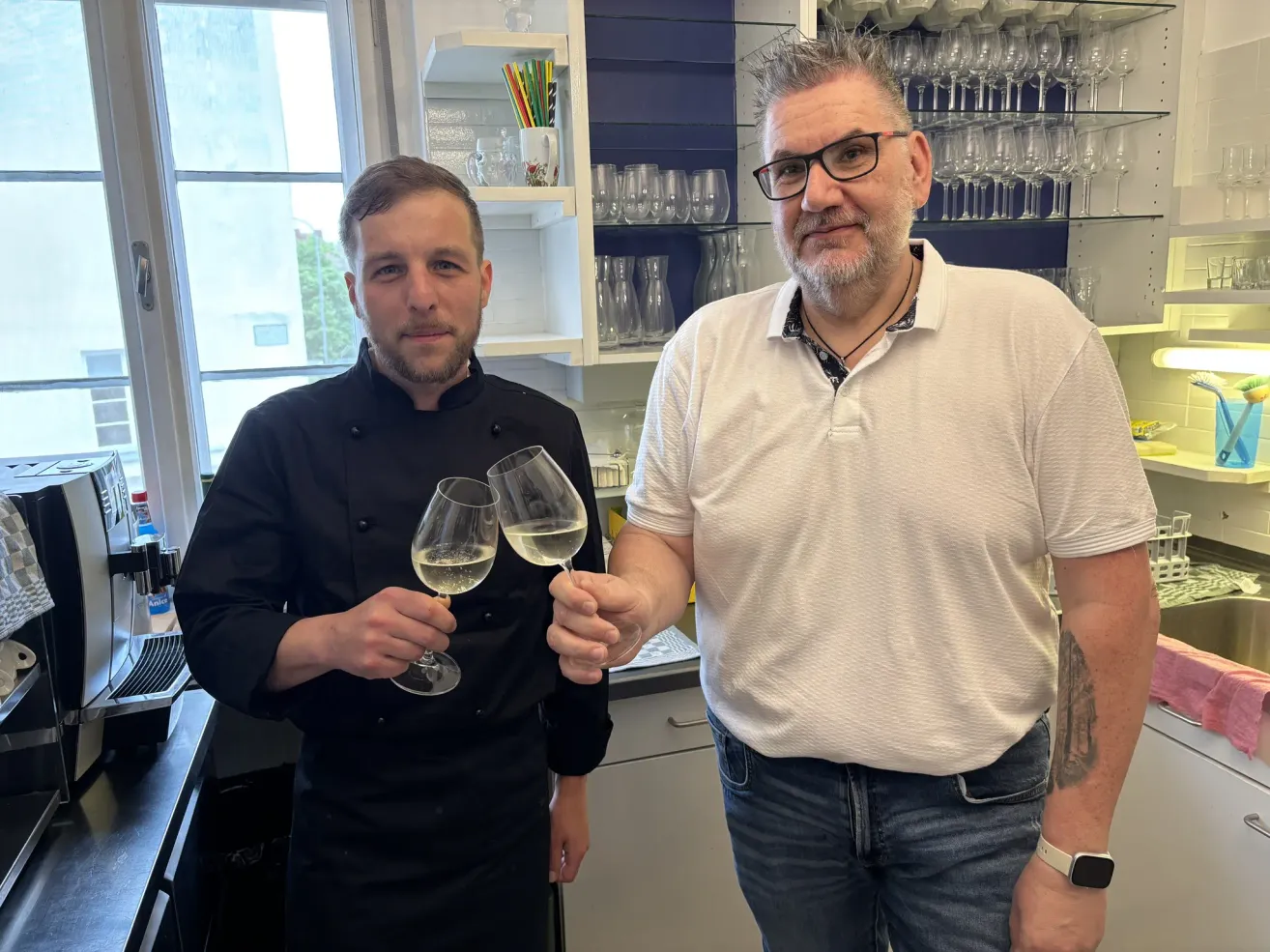 Zwei Männer in einer Küche. Einer in einer Kochuniform hält zwei Weingläser mit Wein. Der andere trägt ein weißes Polohemd und eine Brille. Sie stehen vor Regalen mit Weingläsern.