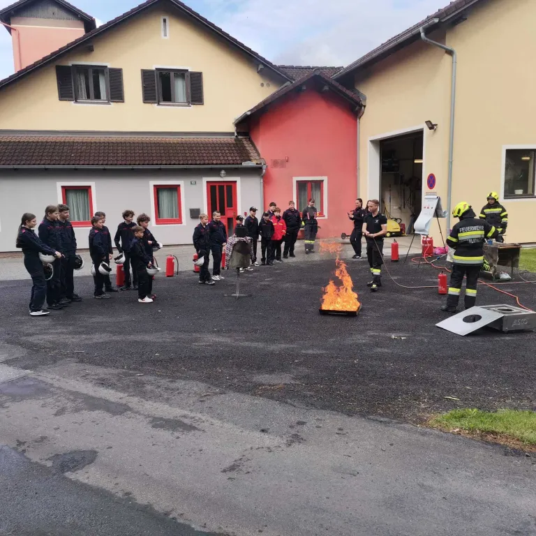 A group of children are standing in line, watching firemen extinguish a small fire on the road in front of a building.