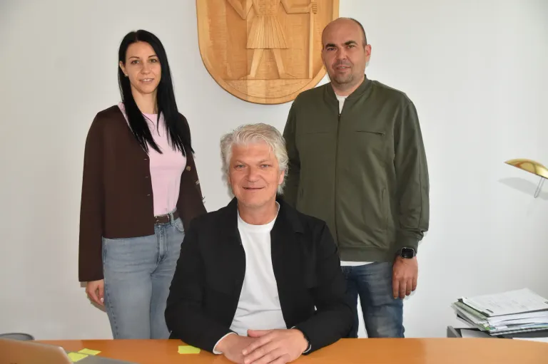 Drei Personen, zwei Frauen und ein Mann, stehen und posieren für ein Foto vor einem hölzernen Wappen an der Wand.