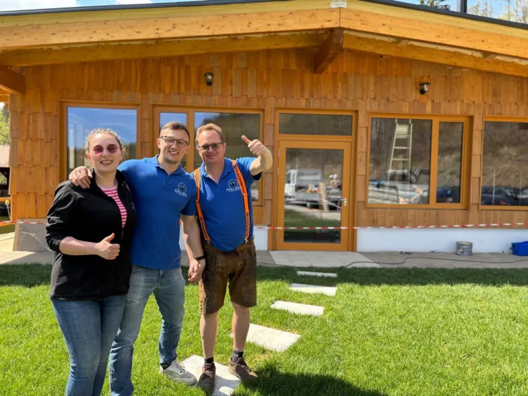Drei Personen stehen vor einem hölzernen Haus mit Glasfenstern. Die beiden Männer tragen blaue Hemden und Hosenträger, während die Frau eine Sonnenbrille und eine schwarze Jacke trägt.