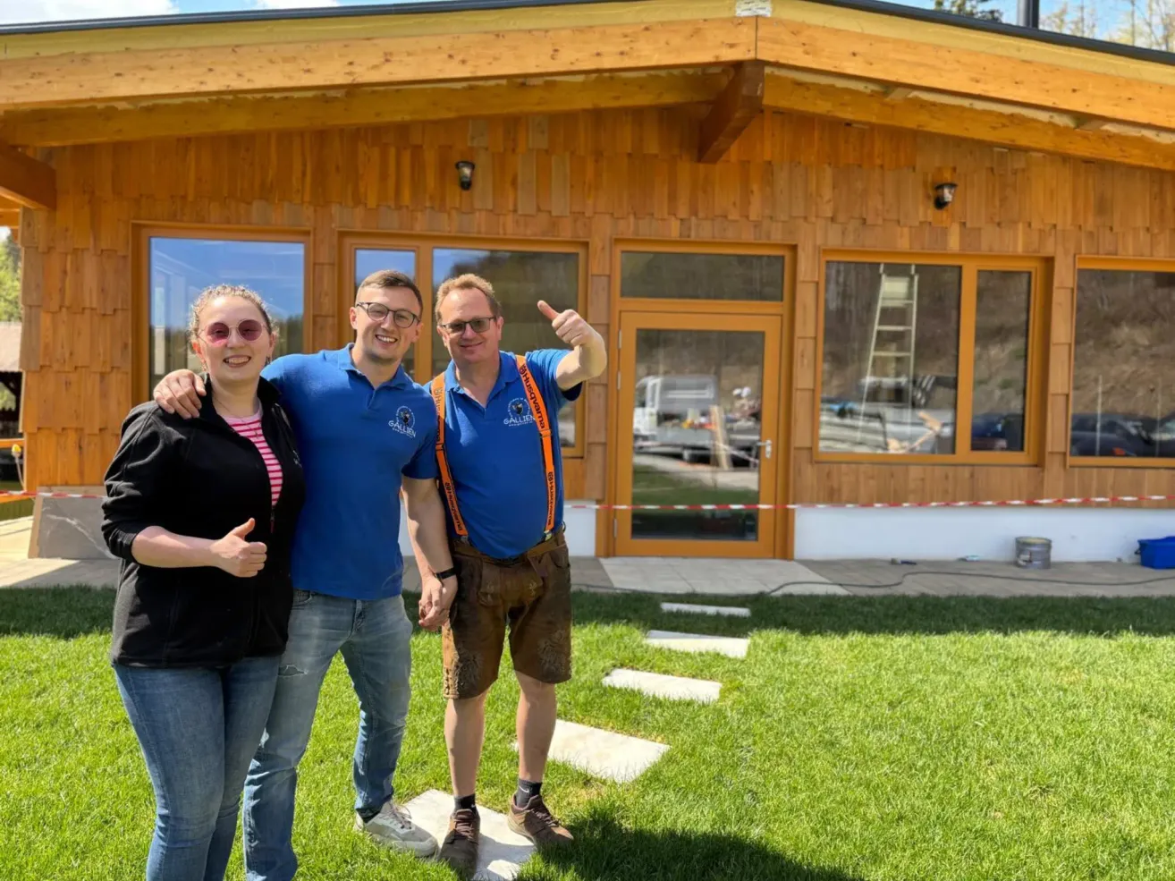 Drei Personen stehen vor einem hölzernen Haus mit Glasfenstern. Die beiden Männer tragen blaue Hemden und Hosenträger, während die Frau eine Sonnenbrille und eine schwarze Jacke trägt.
