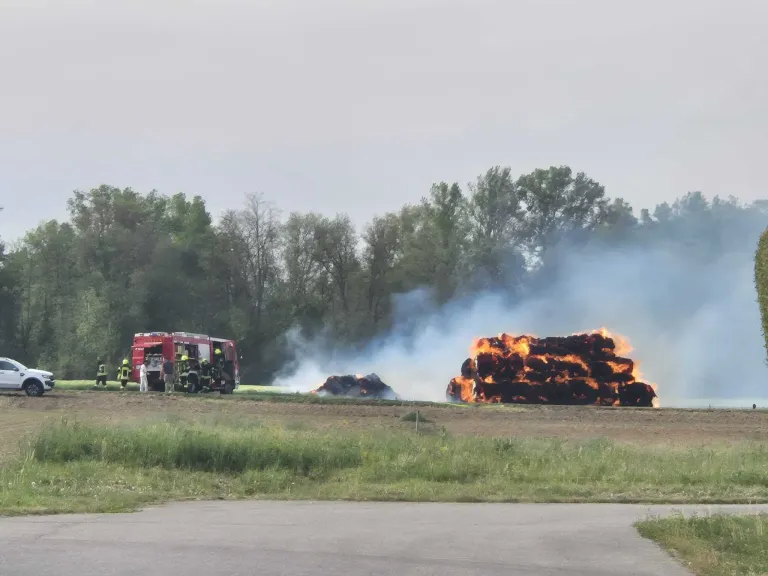 Feuerwehrleute stehen neben einem roten LKW, während sie versuchen, einen großen brennenden Heuhaufen auf einem Feld zu löschen.