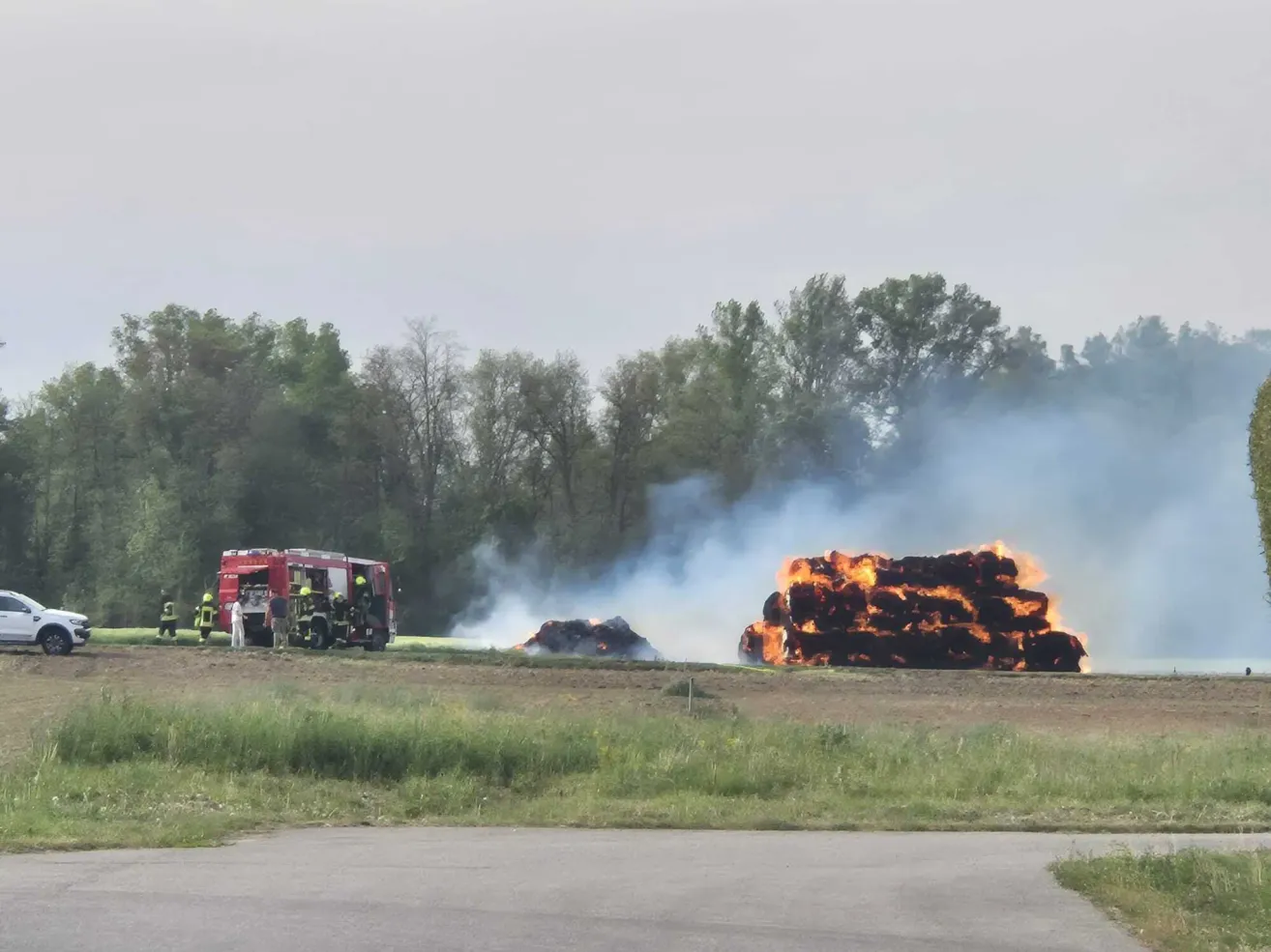 Feuerwehrleute stehen neben einem roten LKW, während sie versuchen, einen großen brennenden Heuhaufen auf einem Feld zu löschen.