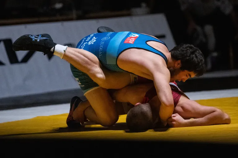 Ein Ringer, der ein blaues Singlet mit einem roten und weißen Logo auf dem Rücken trägt, ringt auf einer gelben Matte mit einem Gegner.
