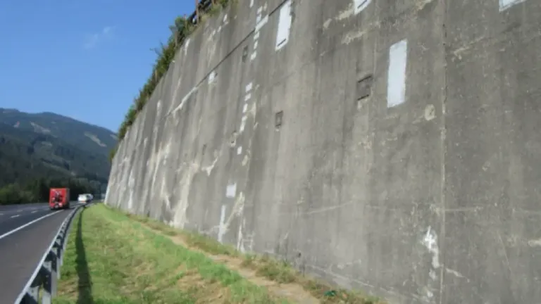 A highway with a concrete wall beside it. A red truck and a white car are driving on it. There are plants on top of the wall.