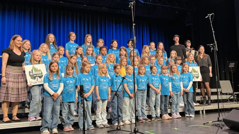 Ein Chor von Kindern in blauen Shirts mit Logos steht auf der Bühne, begleitet von einem Mann in Schwarz. Sie alle schauen in die gleiche Richtung, wahrscheinlich bei einer Veranstaltung.