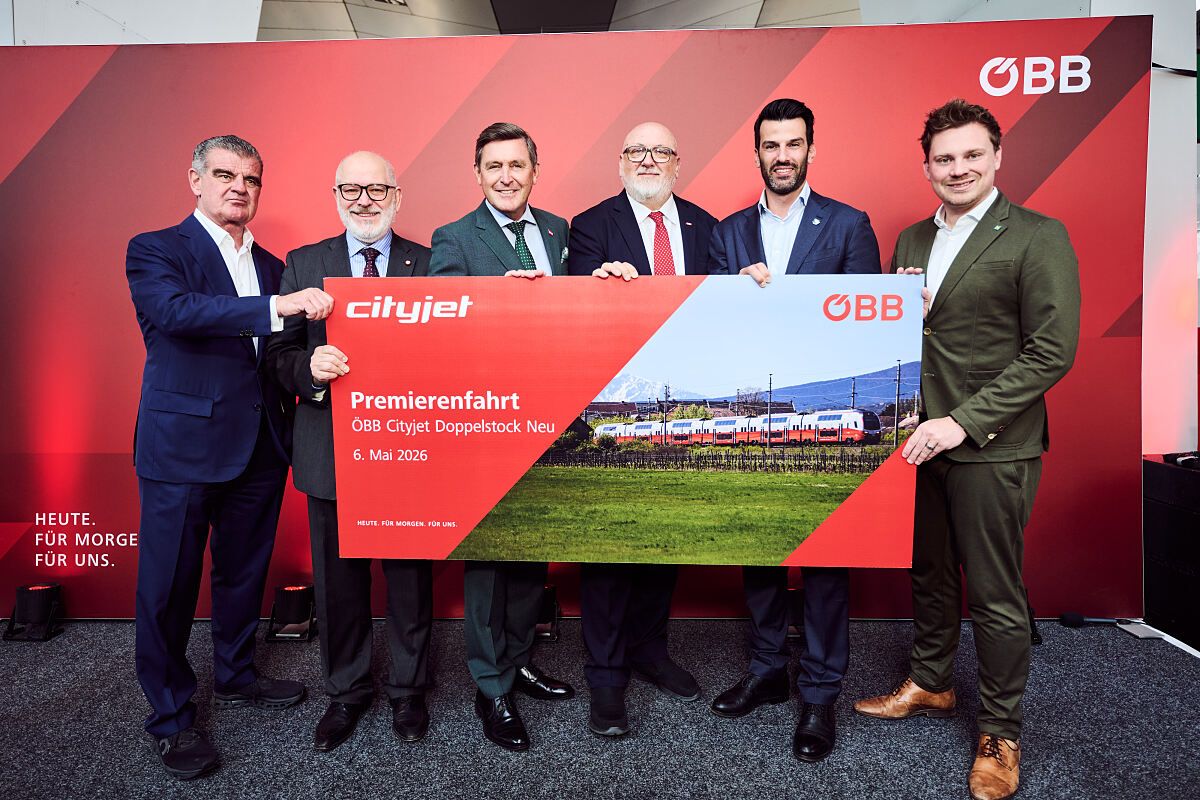 Five men in formal attire stand in front of a red backdrop, holding a red sign that reads 'Cityjet Premierefahrt OBB Cityjet Doppelstock Neu 6. Mai 2026'. The sign has a picture of a train.
