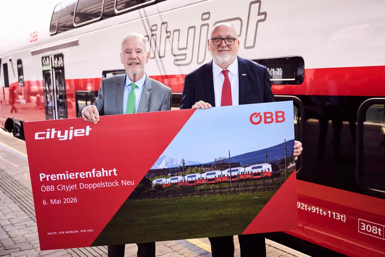 Two men holding a large sign with a train image in front of a train, one man wears a green tie and the other a red tie.