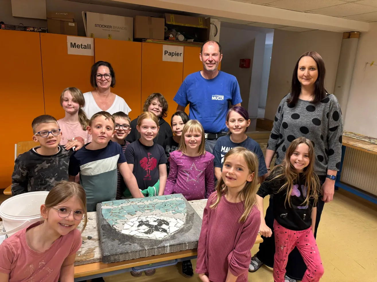 Eine Gruppe von Kindern und Erwachsenen posiert für ein Foto in einem Raum. Sie sind um einen Tisch mit einem Mosaik versammelt. Zwei Frauen und ein Mann stehen hinter den Kindern. Der Raum hat Schränke mit Schildern, die Metall und Papier bedeuten.