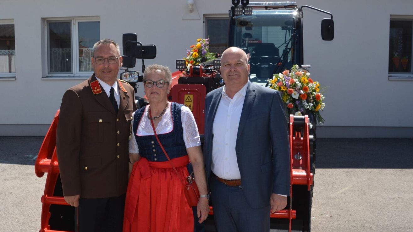 Three individuals stand in front of a red tractor. A woman in a traditional dress, and two men, one in military uniform and the other in a suit.