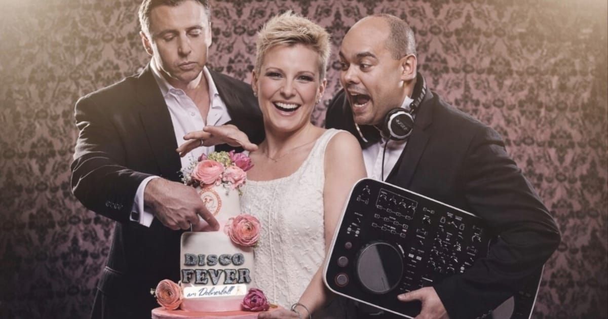 A smiling bride holds a cake labeled 'Disco Fever' while two men, one holding a DJ mixer, surround her. The scene is set against a floral wallpaper.