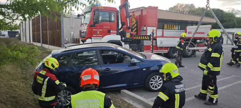 Firefighters are assisting a blue car near a red fire truck with a crane and a building in the background.