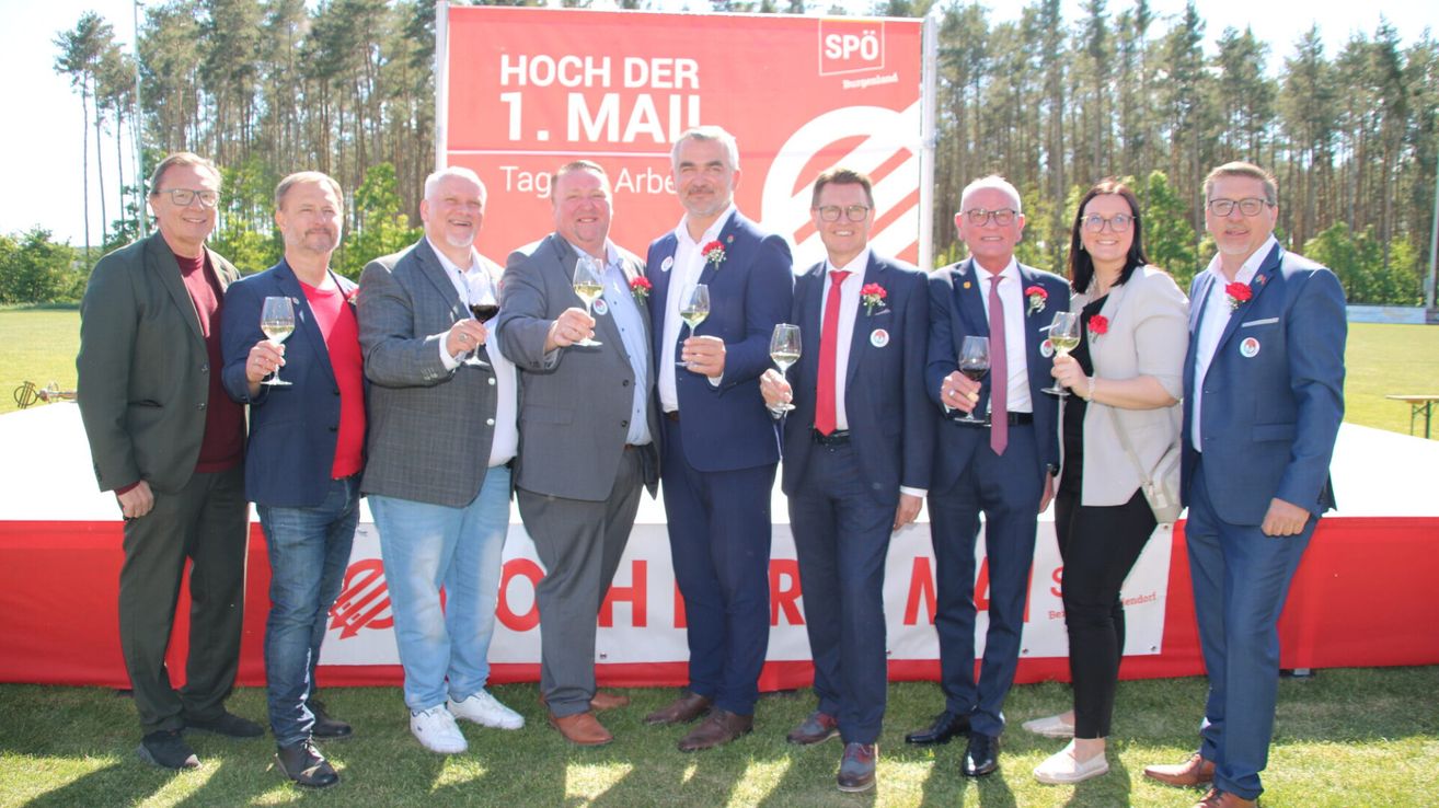 Six men in suits and one woman are standing on a lawn. They all hold wine glasses. Behind them is a banner with the text 'Hoch der 1. Mai'.