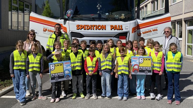 Eine Gruppe von Kindern in gelben Westen steht vor einem Lastwagen mit der Aufschrift 'SPEDITION PALL'. Sie halten Schilder mit der Aufschrift 'Freunde auf der Straße'.